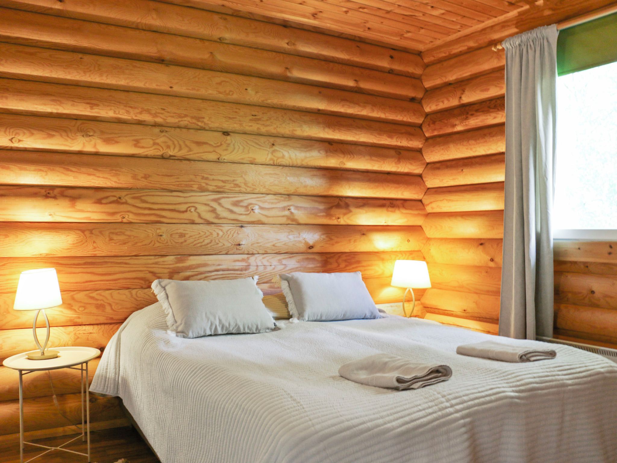 A wooden cabin bedroom with a bed, two bedside lamps, and a window with curtains.