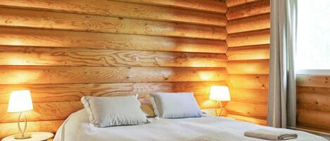 A wooden cabin bedroom with a bed, two bedside lamps, and a window with curtains.