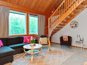 Una sala de estar acogedora con techo de madera, una escalera, un sofá, un sillón, una mesita y vistas a árboles a través de grandes ventanales.