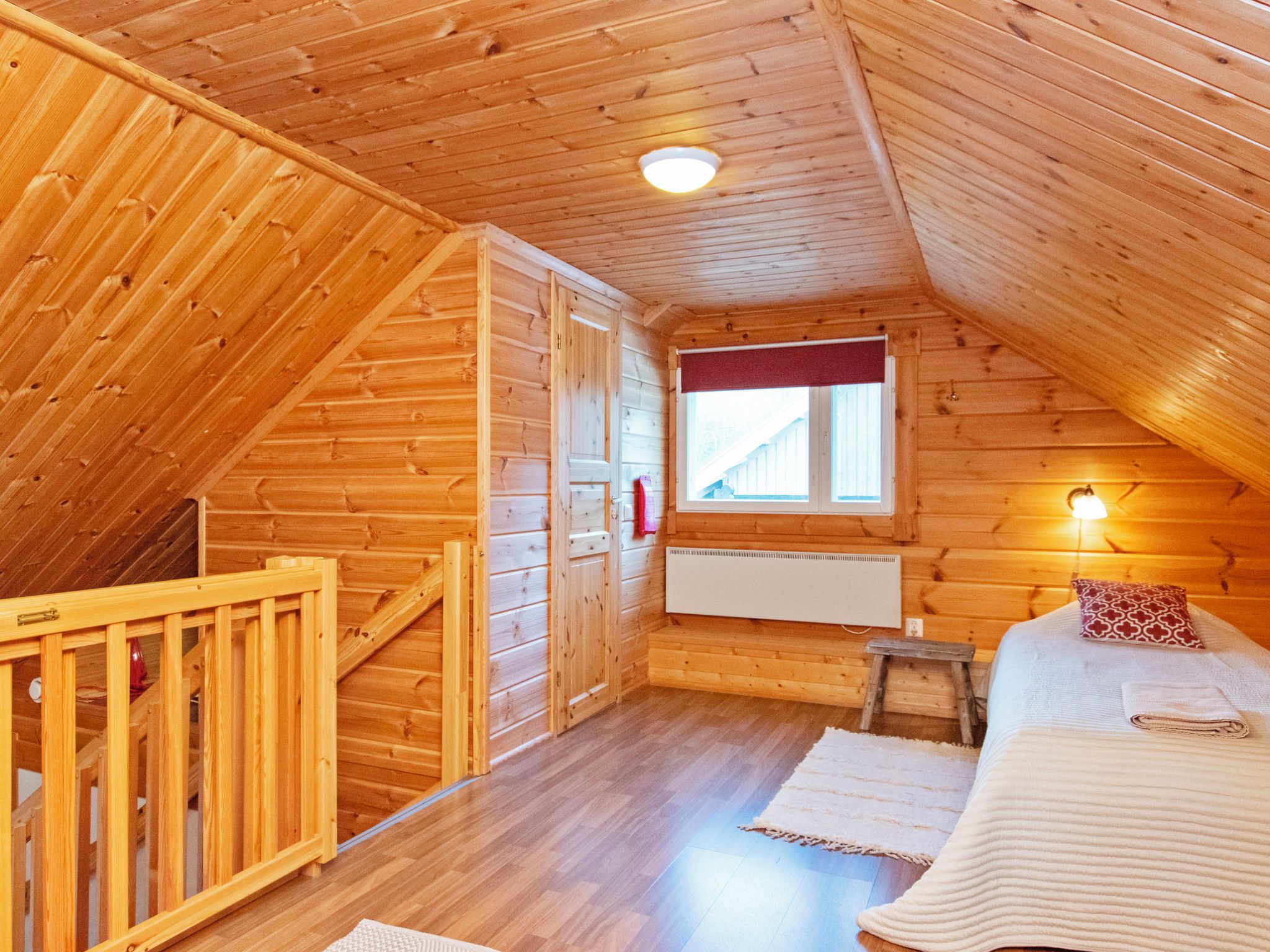 A cozy attic bedroom with wooden walls, a sloped ceiling, a window with a red curtain, a bed with a white blanket, and a small stool.