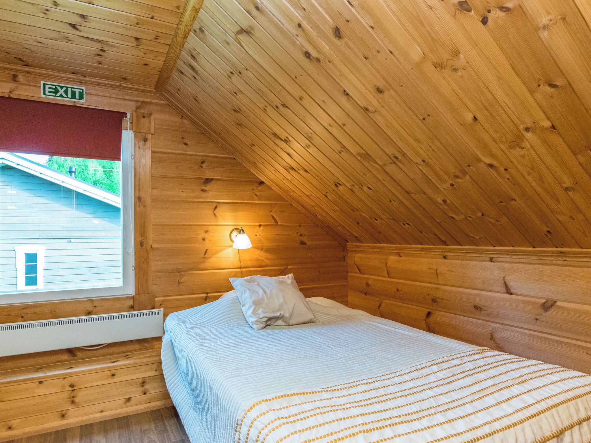 A single bed in a cabin with a sloped ceiling and an exit sign above the door.
