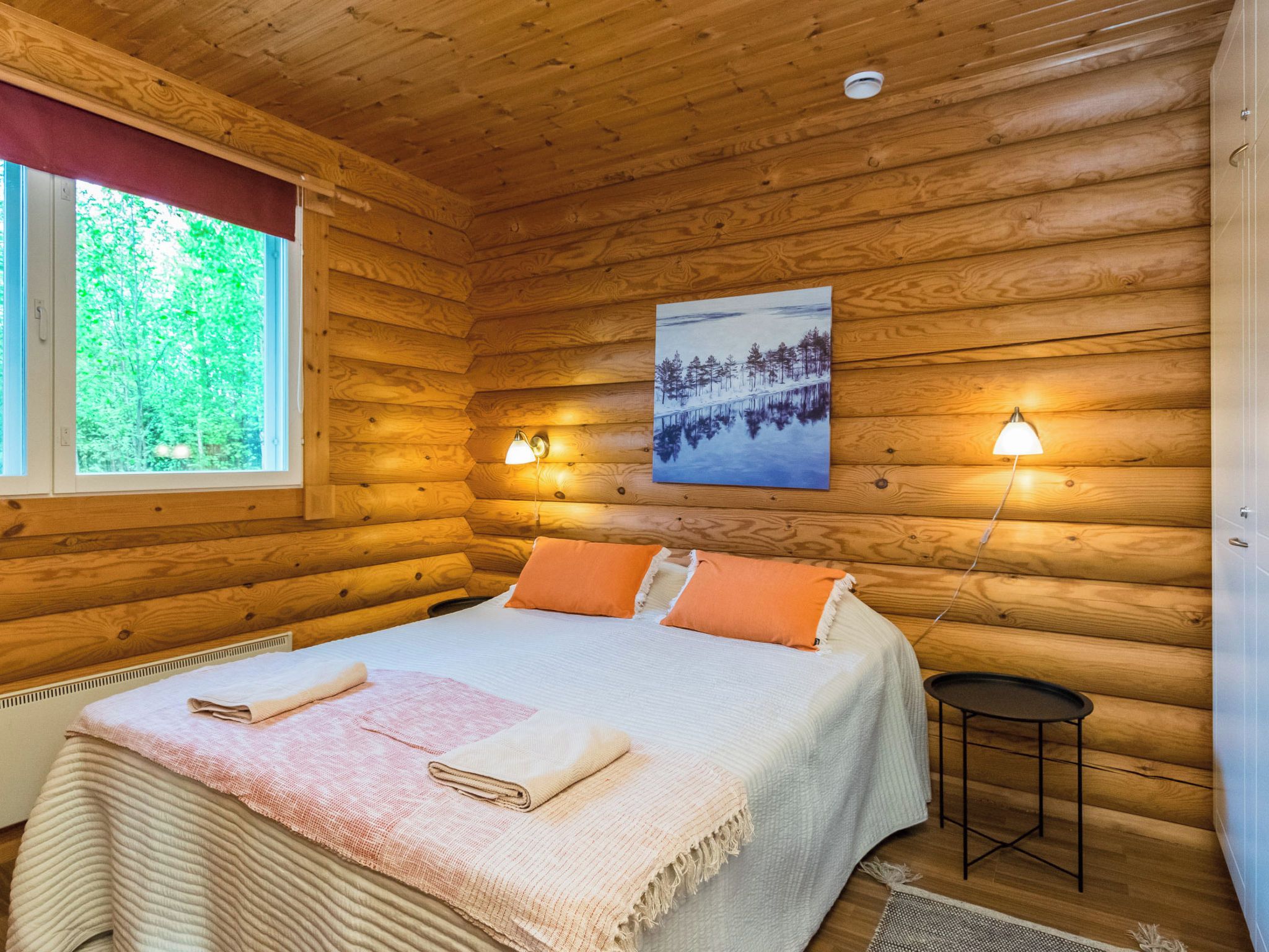 En stuga med träpanel, ett rum med en säng, två sänglampor, en tavla och ett fönster med utsikt över träd.