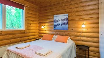 En stuga med träpanel, ett rum med en säng, två sänglampor, en tavla och ett fönster med utsikt över träd.