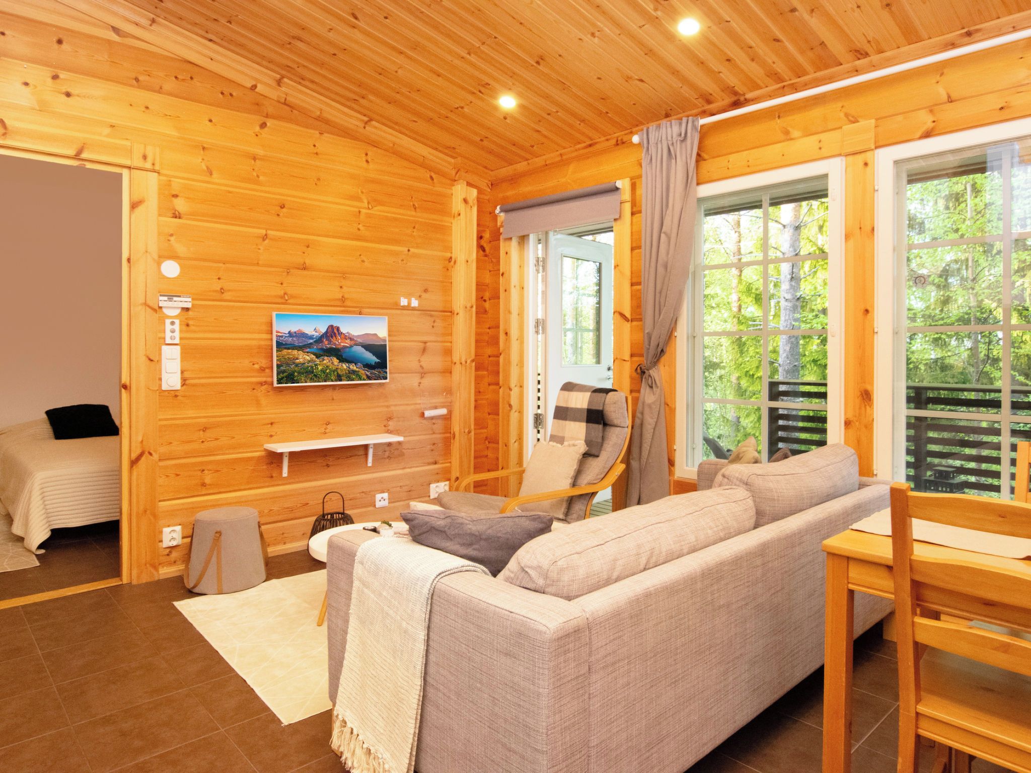 A wooden cabin interior with a sofa, a chair, a small table, a bed, and a window with a view of trees.