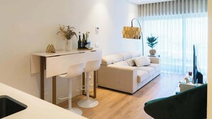 A modern living room with a sofa, a dining area with bar stools, and a large window with sheer curtains.