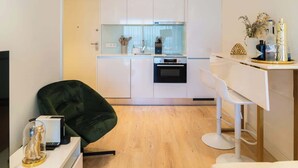 A modern kitchen with white cabinets, a built-in oven, and a glass backsplash. There is a bar area with a white chair and a small table.