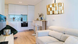 A modern living room with a white sofa, a dining table with chairs, and a kitchen area with white cabinets and a built-in oven.