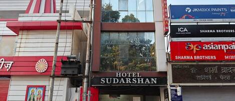 A hotel named 'Hotel Sudarshan' with a prominent sign, a red and white facade, and a large window display.