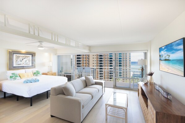 A spacious living room with a large bed, a sofa, and a coffee table. There is a balcony with a view of the city and a painting of a beach on the wall.