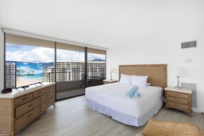 Quarto de hotel com uma grande janela, cama, cômoda, mesa de cabeceira e uma cadeira de vime.