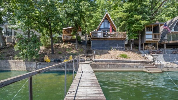A wooden dock leading to a house with a deck, surrounded by trees and a body of water.