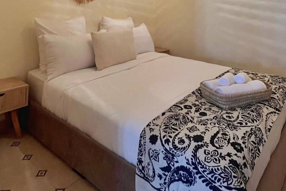 A neatly made bed with white linens, a patterned throw blanket, and a bedside table with a lamp.