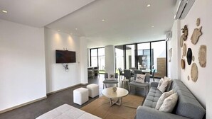 Amplio salón con sofá gris, otomanas blancas y una mesa de centro redonda. Hay un televisor de pantalla plana montado en la pared, y la habitación tiene ventanas grandes que proporcionan luz natural.