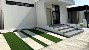 Una casa moderna de dos pisos con techo plano, ventanas amplias y un pequeño jardín delantero con césped artificial y escalones que conducen a la entrada.