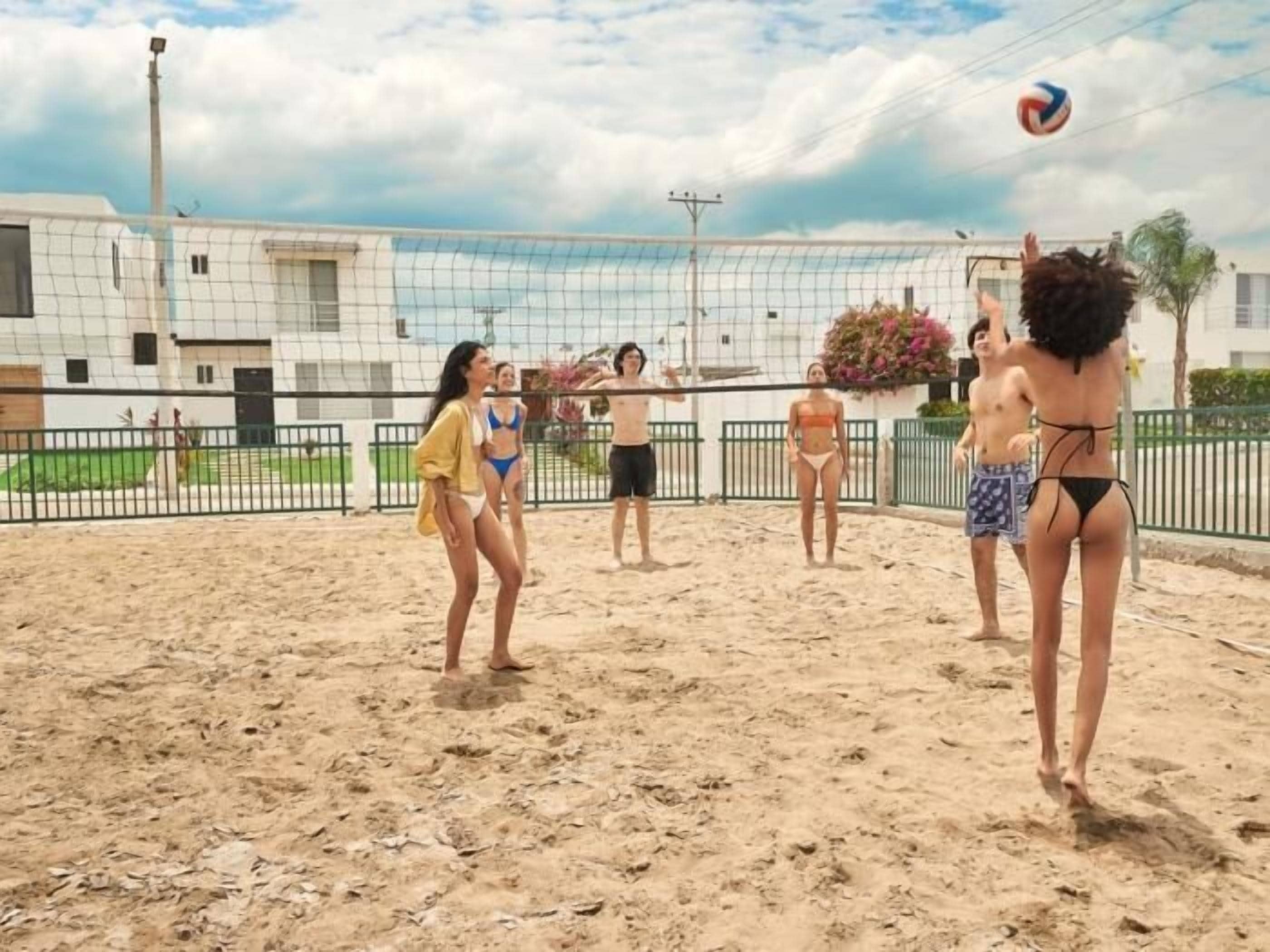 Un match de volleyball se déroule sur un terrain de sable avec un filet, entouré d’une clôture et avec des bâtiments en arrière-plan.