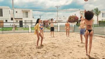 Un match de volleyball se déroule sur un terrain de sable avec un filet, entouré d’une clôture et avec des bâtiments en arrière-plan.