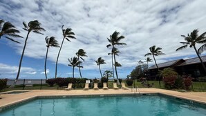 Une piscine entourée de chaises longues, de palmiers et d’une clôture, sous un ciel partiellement nuageux.