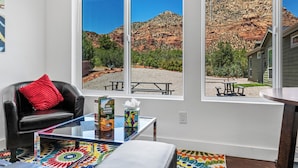 Una habitación con un gran ventanal con vistas a un paisaje montañoso, un sillón de cuero con un cojín rojo y una mesa de centro de vidrio con una alfombra colorida.