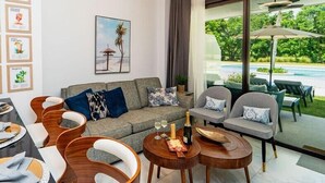 A modern living room with a grey sofa, wooden chairs, and a dining table set with plates and glasses. A painting of a beach scene hangs on the wall, and a window offers a view of a pool and greenery.