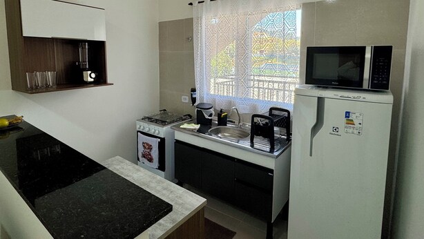 A compact kitchen with a gas stove, a microwave, and a refrigerator. There is a window with patterned curtains, and a shelf with glasses and a blender. - Cozy house in Polvilho Cajamar, just a few minutes from Rod. Anhanguera (Jardim Santana Cajamar)