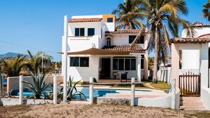 Una casa de dues plantes amb piscina, envoltada de palmeres i un cel blau clar.