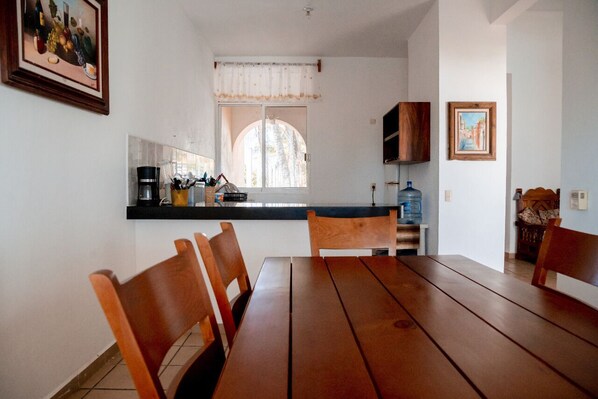 Un coin repas avec une table et des chaises en bois, une cuisine avec une fenêtre et un tableau accroché au mur.