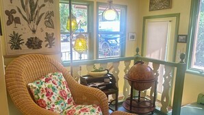 Een rieten stoel met bloemenkussens, een hanglamp en een ingelijste botanische print aan de muur.