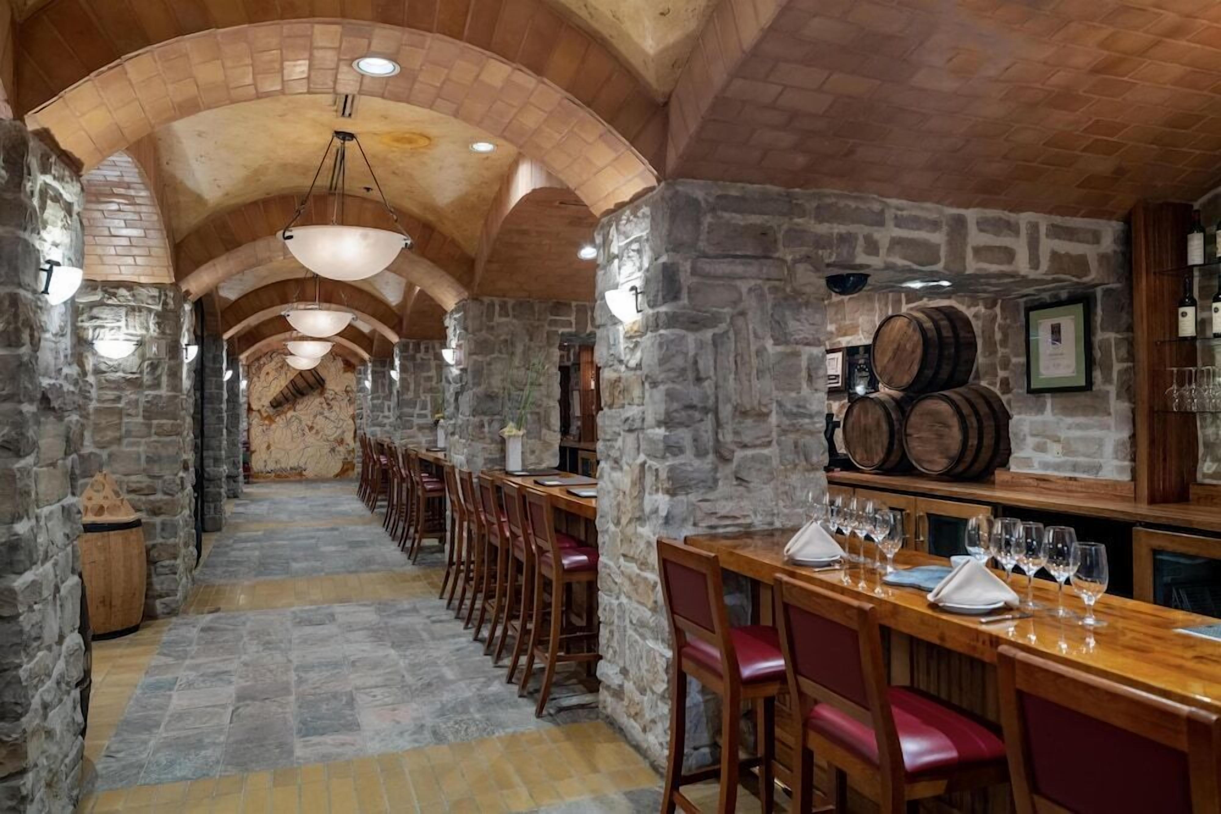 Un espace bar aux murs de pierre, avec des tables et des chaises en bois, et un long comptoir où sont exposés des verres à vin.