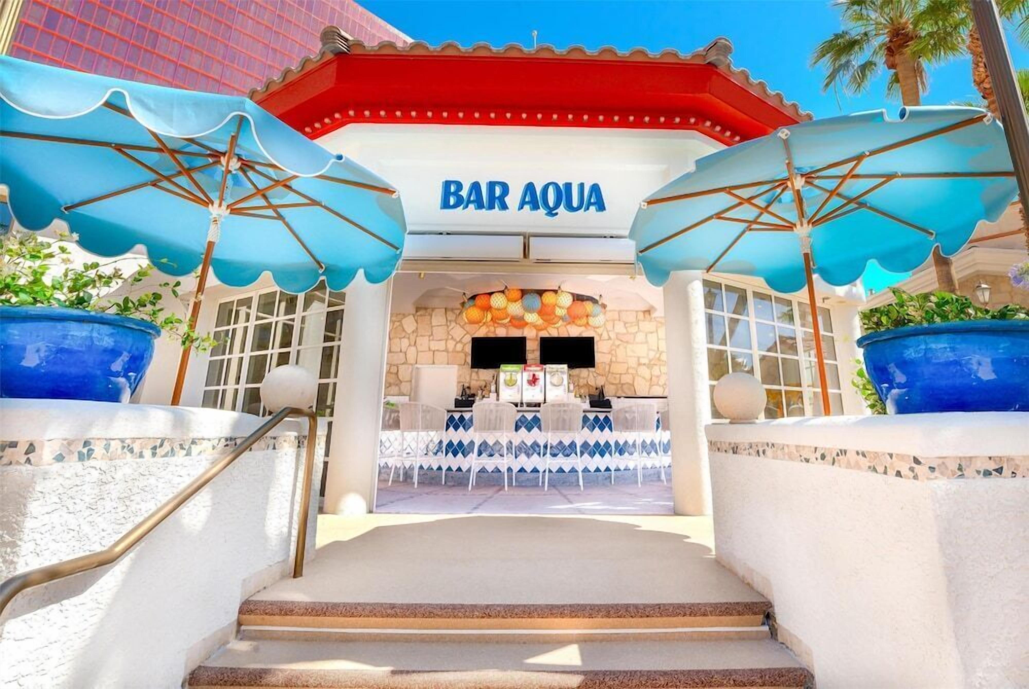 L’entrée du Bar Aqua se distingue par sa façade en pierre, ses parasols bleus et un escalier menant à l’étage supérieur.