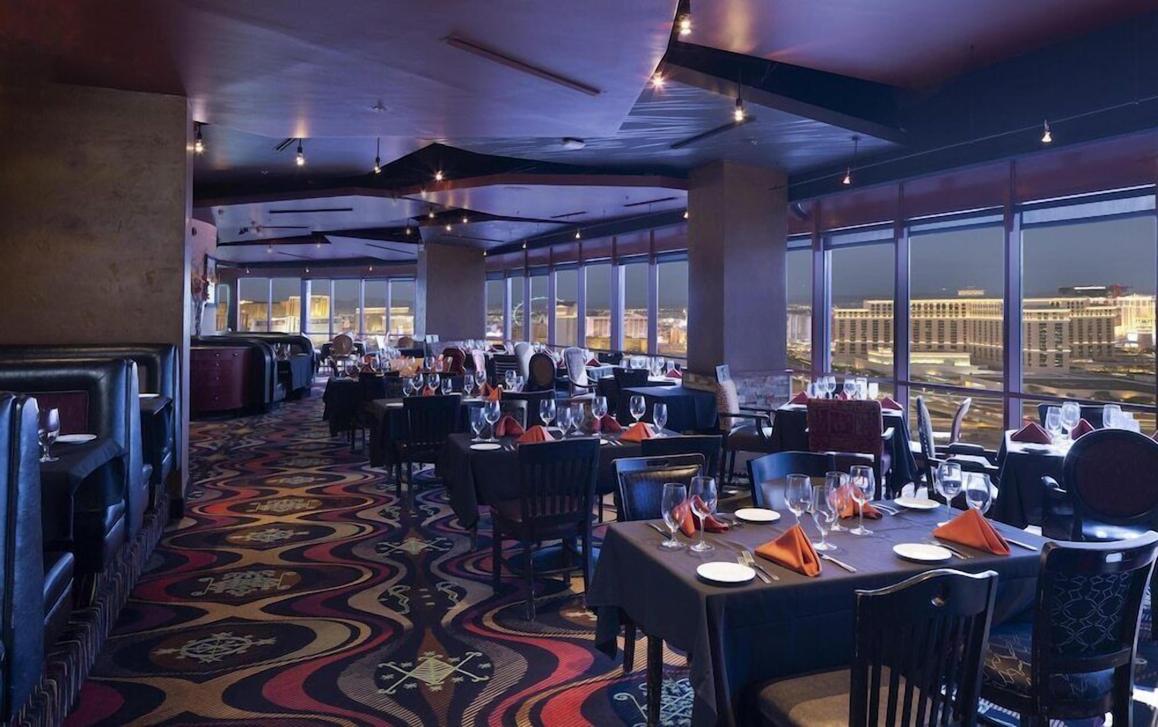 Un restaurante con mesas dispuestas para comer, grandes ventanales que ofrecen vistas a la ciudad y una alfombra estampada.
