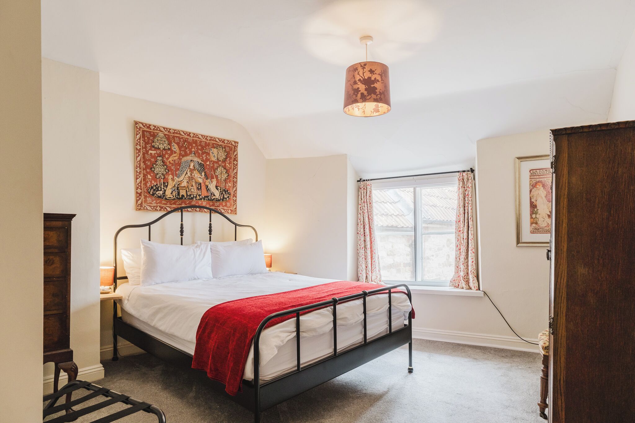 A bedroom with a bed, a nightstand, a lamp, a window with curtains, and a tapestry on the wall.