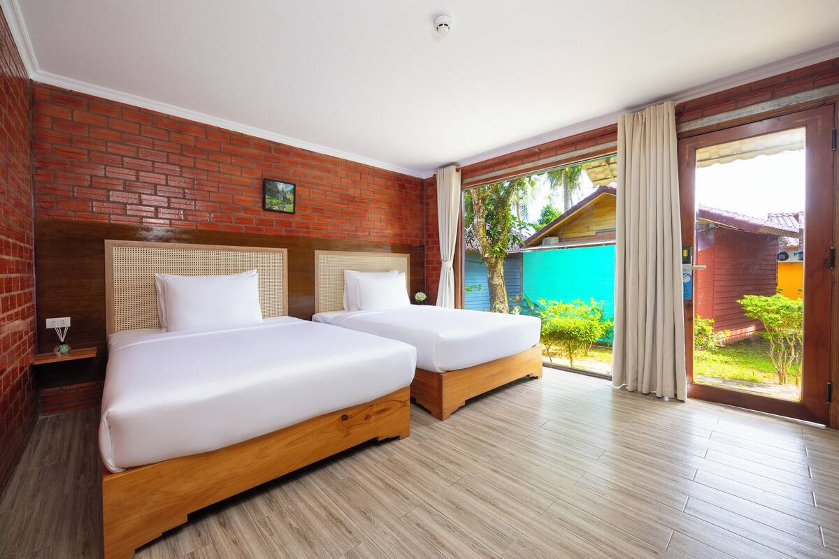 A hotel room with two wooden beds, a brick wall, and a large sliding glass door leading to a balcony with a view of a blue building and greenery.