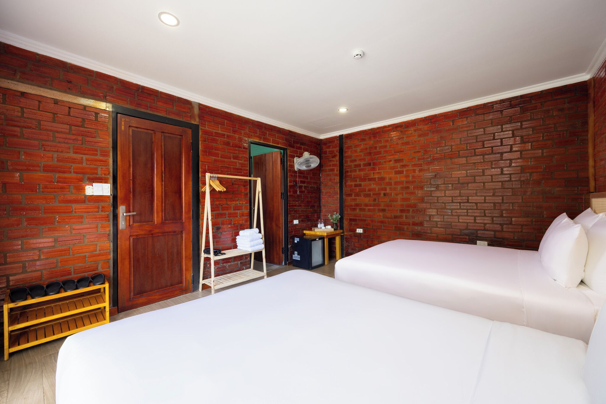 A hotel room with a brick wall, a wooden door, a bed, a small desk, and a shelf with towels.