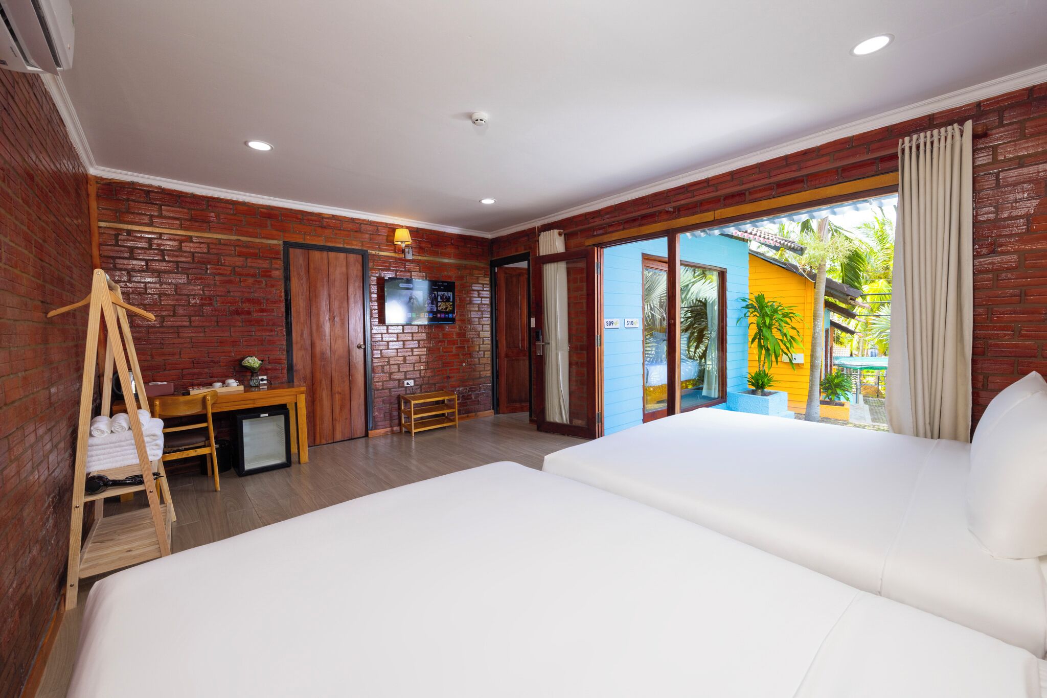 A hotel room with two beds, a desk, a TV, and a balcony with a view of the outside.