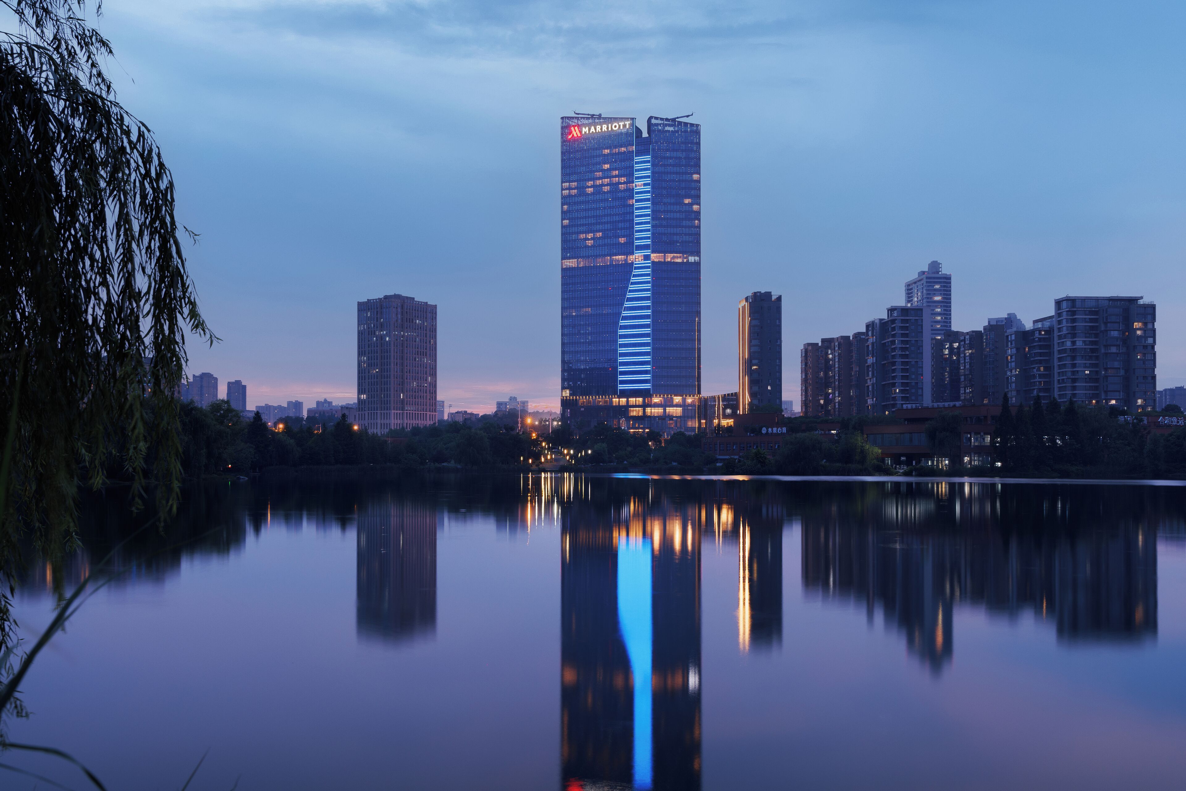 A high-rise hotel with a modern design, situated by a calm body of water, reflecting the building and the surrounding cityscape.