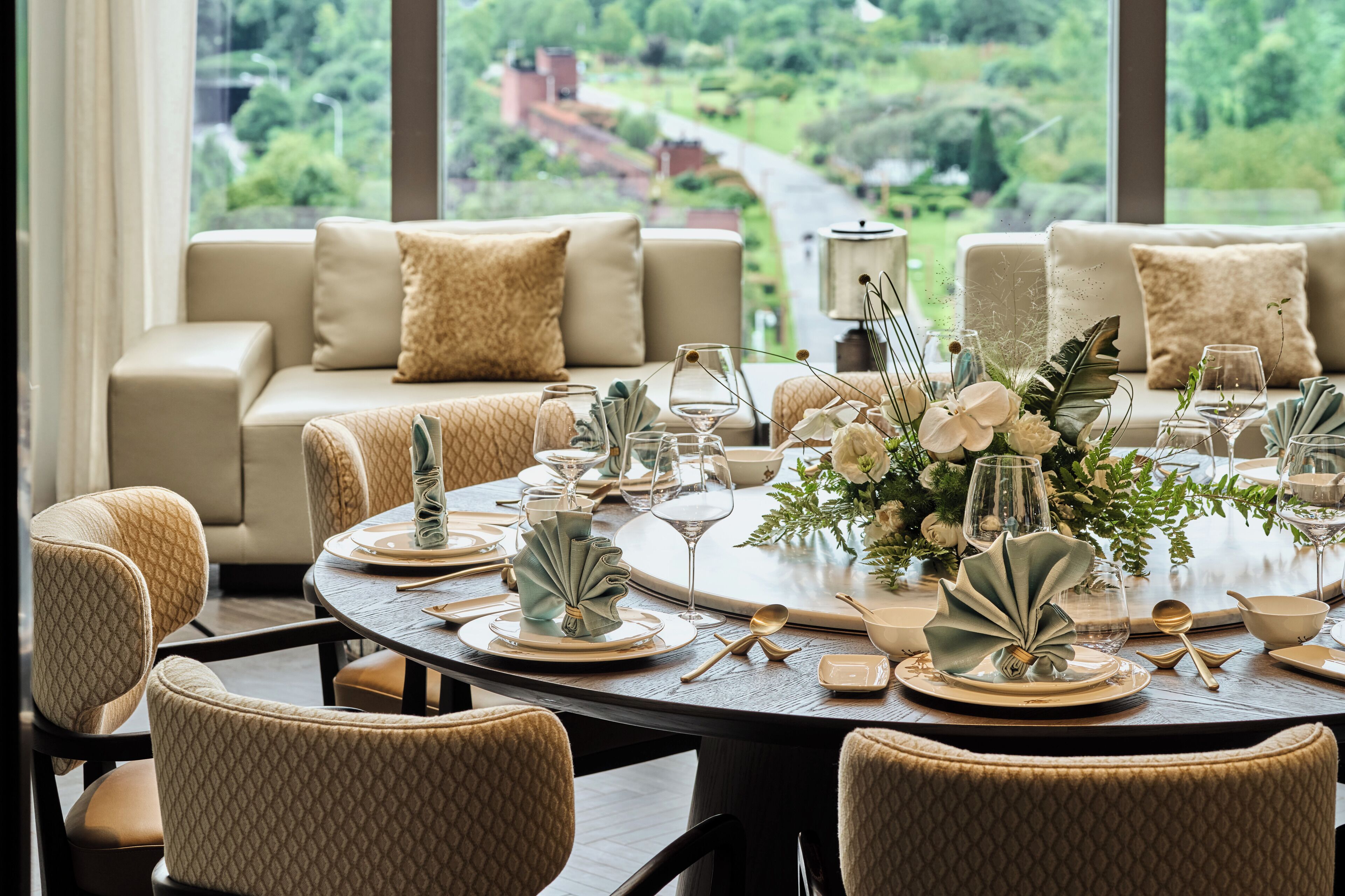 A dining area with a round table set for six, surrounded by beige upholstered chairs. A sofa with beige cushions is visible in the background, and the room has large windows offering a view of a green landscape.