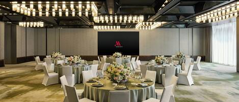 Sala de conferências com mesas redondas, cadeiras brancas e uma grande tela exibindo o logotipo da Marriott.