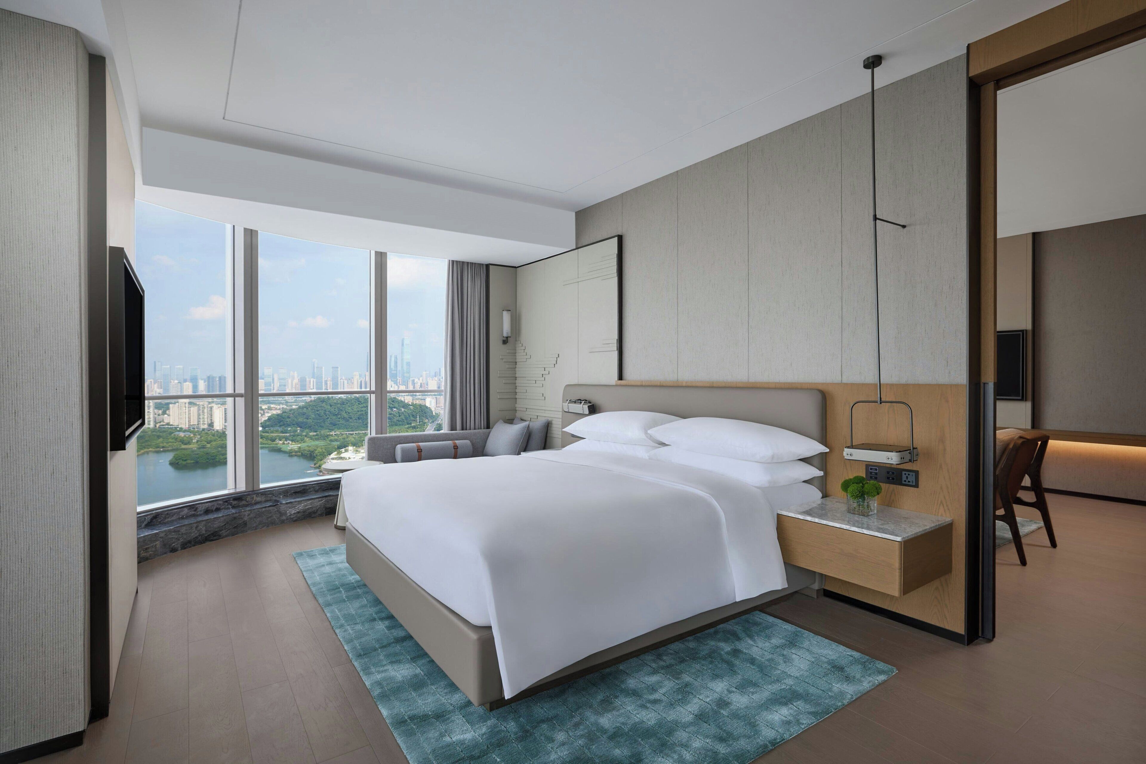 A modern hotel room with a large bed, a bedside table with a lamp, a view of the city, and a large window.