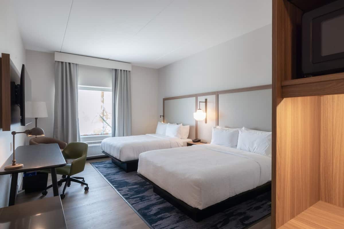 Une chambre d’hôtel avec deux lits, un bureau, une chaise, une lampe et une grande fenêtre avec des rideaux.
