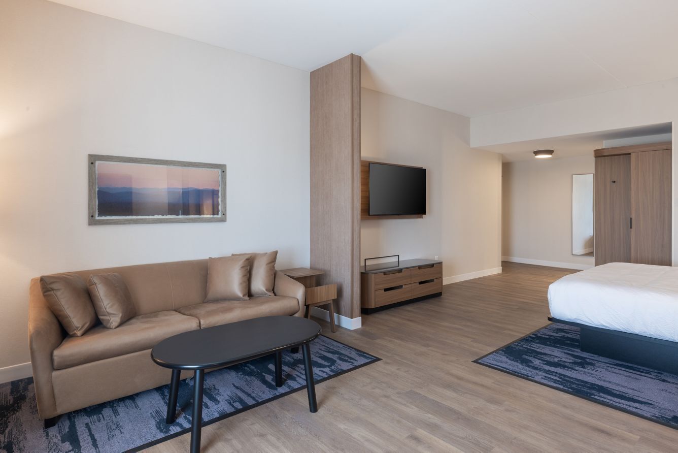 A modern hotel room with a sofa, a TV mounted on the wall, a bed, and a small table.