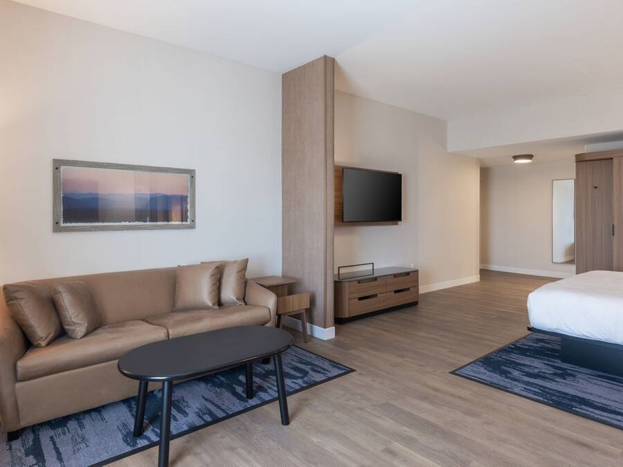 A modern hotel room with a sofa, a TV mounted on the wall, a bed, and a small table.