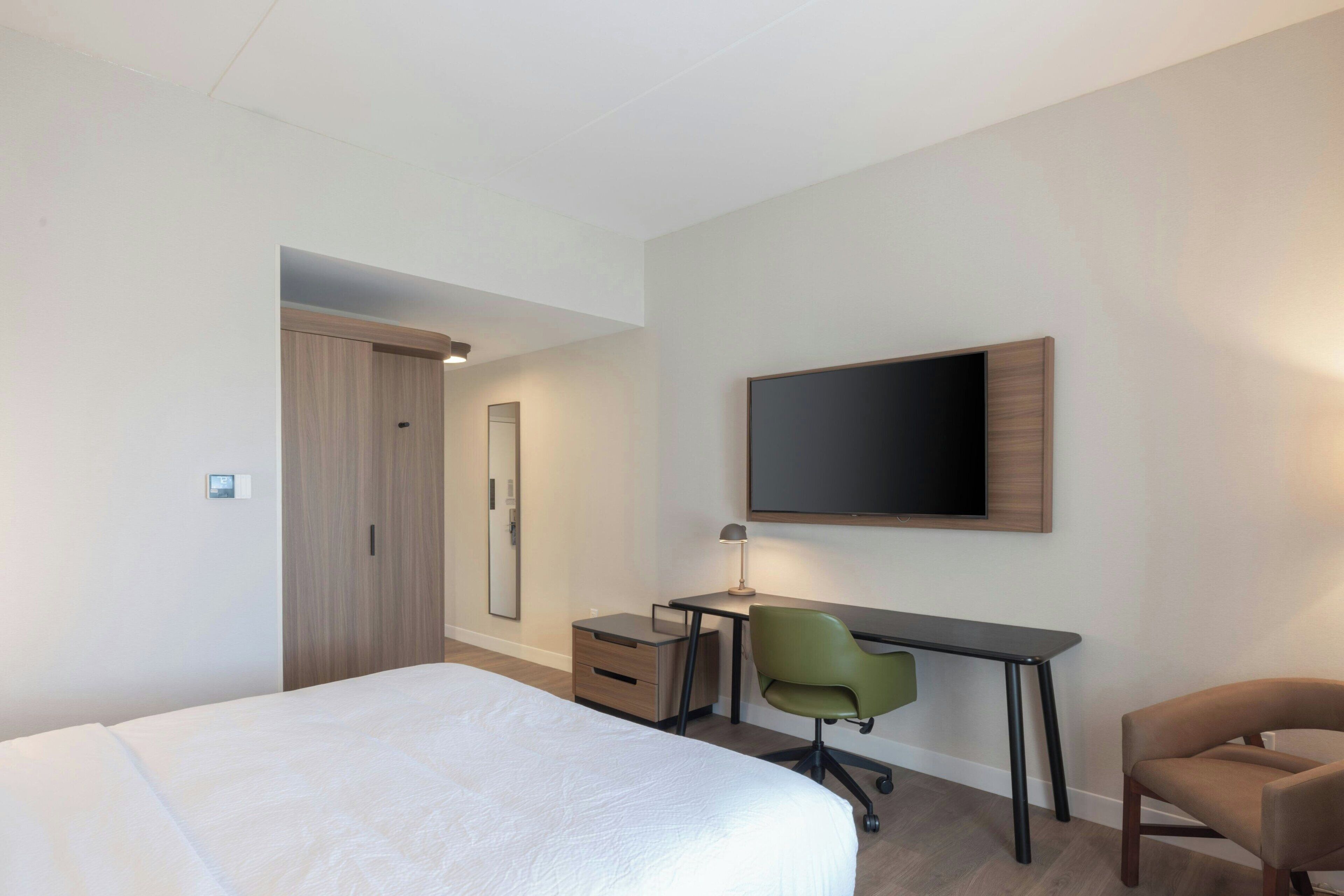 Ein modernes Hotelzimmer mit einem großen Bett, einem Schreibtisch mit Stuhl, einem Flachbildfernseher und einem Kleiderschrank.
