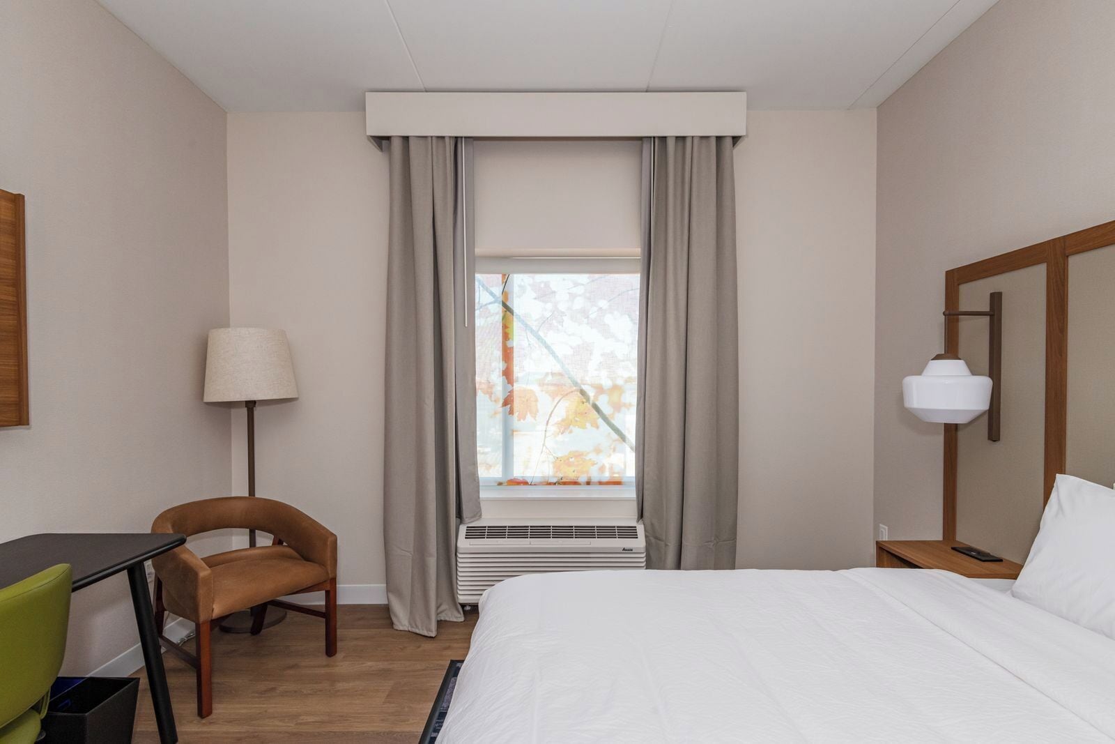 A hotel room with a bed, a desk, a chair, a lamp, and a window with curtains.