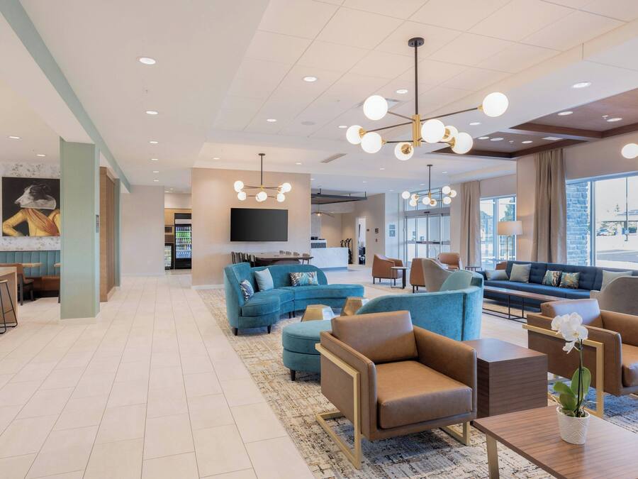 A modern hotel lobby with a blue sofa, brown armchairs, a coffee table, and a large TV screen.