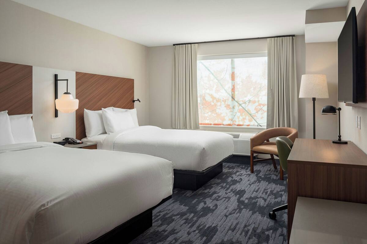 A hotel room with two beds, a desk, a chair, a TV, and a window with curtains.