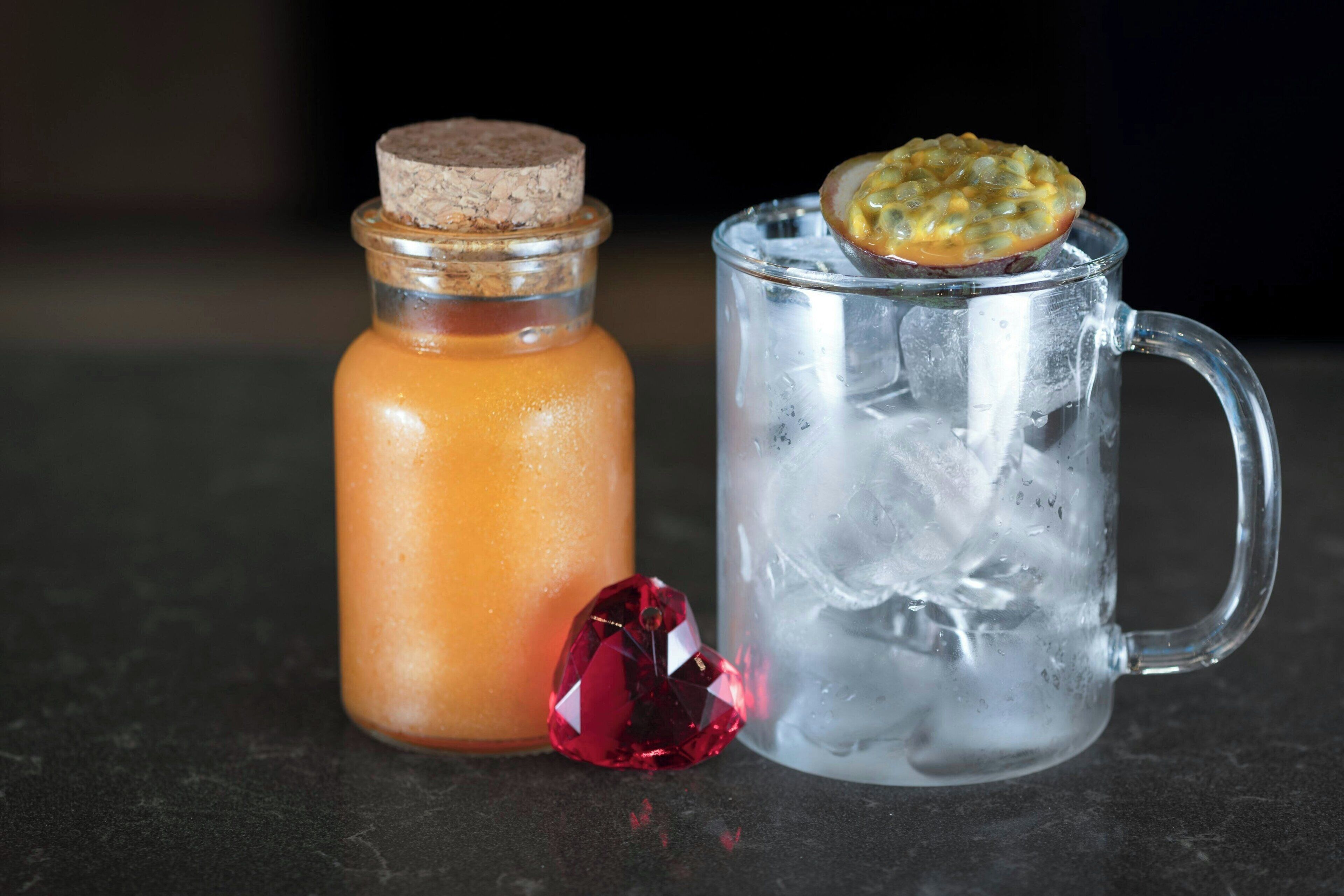 A clear glass mug with ice and a slice of passion fruit on top, next to a glass bottle with a cork stopper.