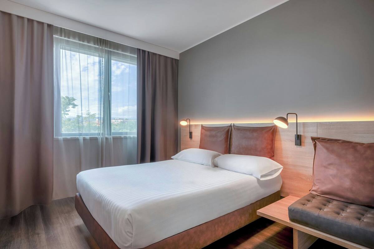A modern hotel room with a large bed, a bedside table, a sofa, and a window with curtains.
