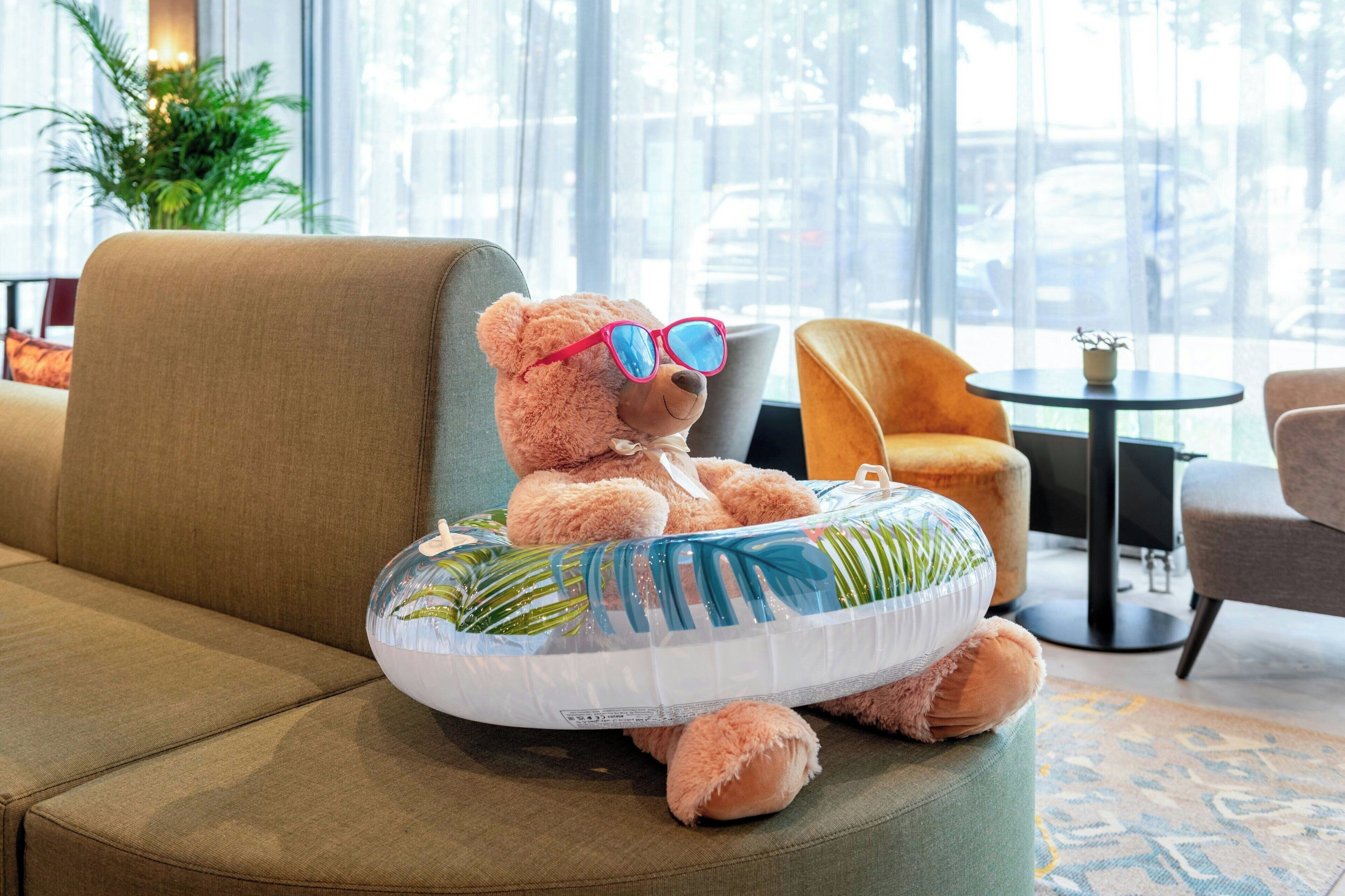 Ein weicher Teddybär mit Sonnenbrille sitzt auf einem runden, gepolsterten Hocker in einem hellen Raum mit einer Couch und einem kleinen Tisch.