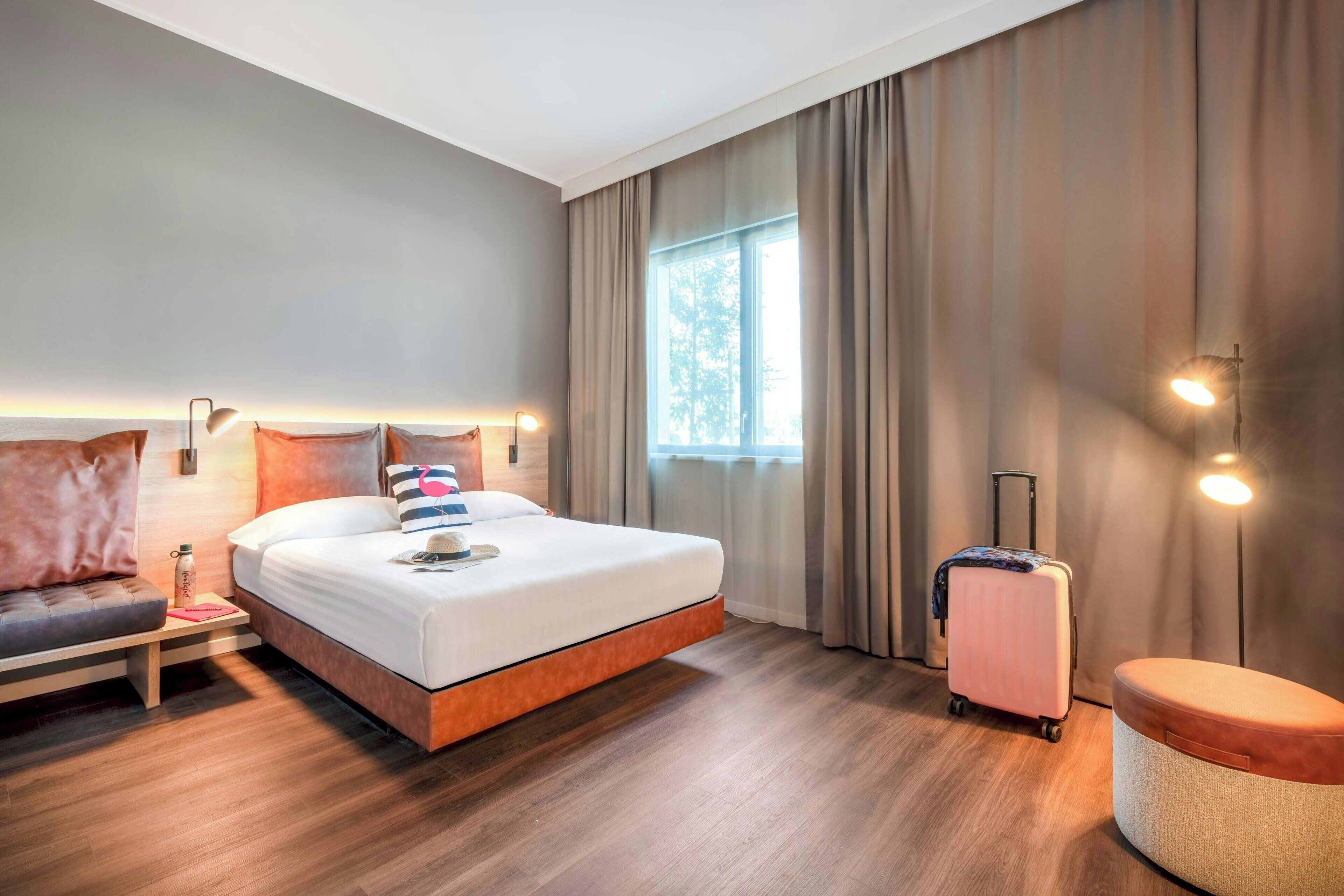 A hotel room with a bed, a desk, a chair, a suitcase, and a window with curtains.