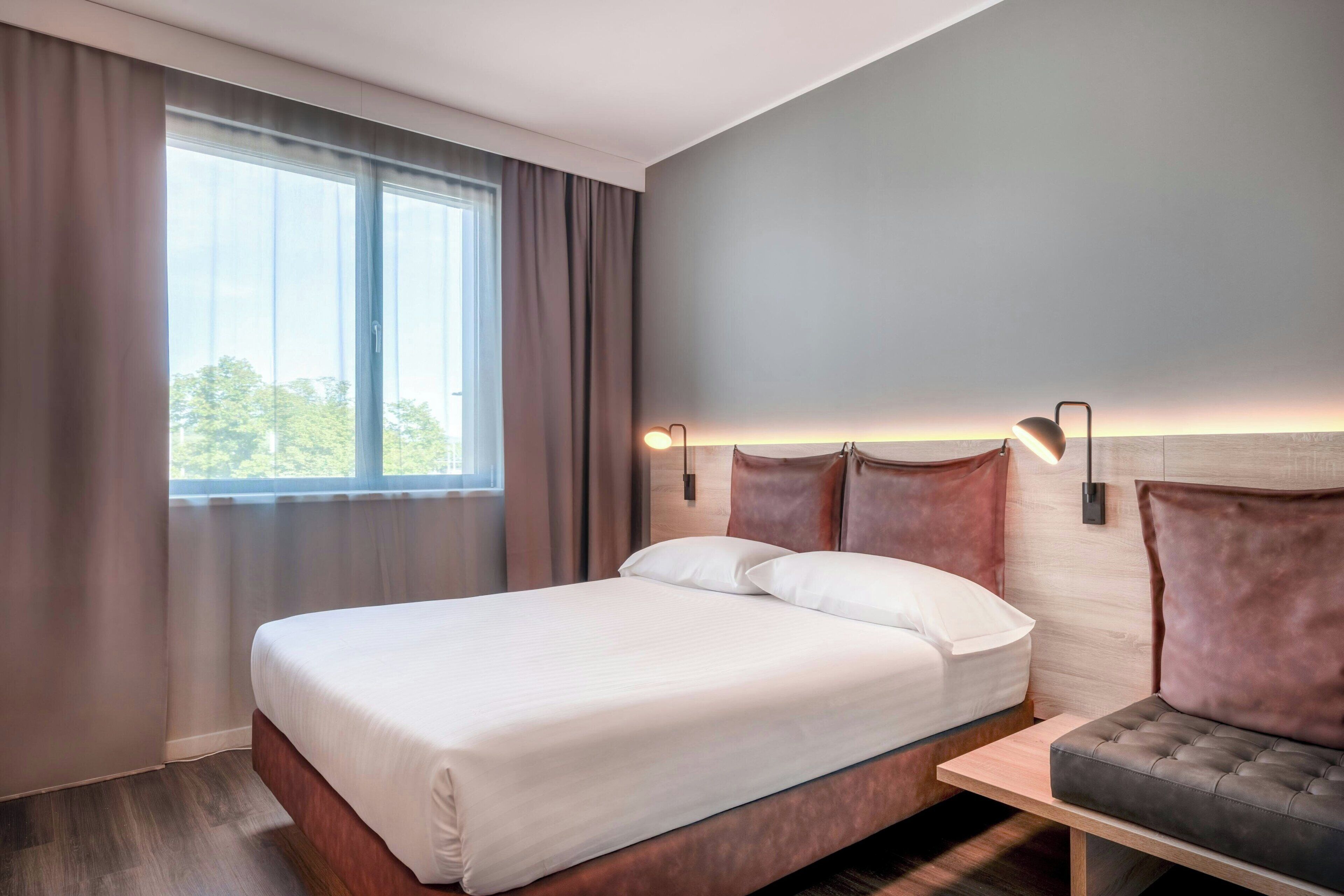 A modern hotel room with a large bed, a bedside table, and a view of the outdoors through a window.